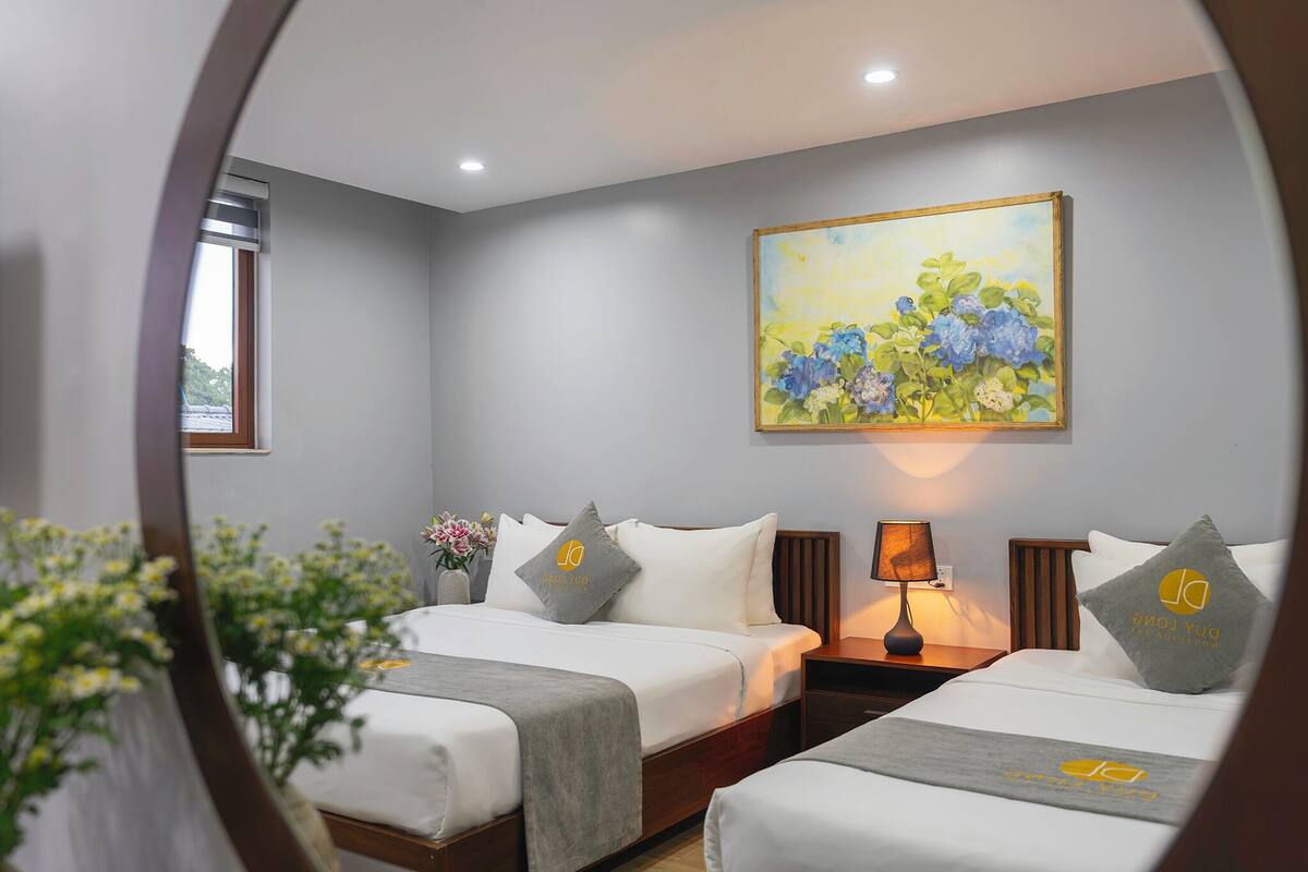 Ein Hotelzimmer mit zwei Betten, einem Blumengemälde an der Wand, einem Nachttisch mit Lampe und einem Fenster mit Blick.