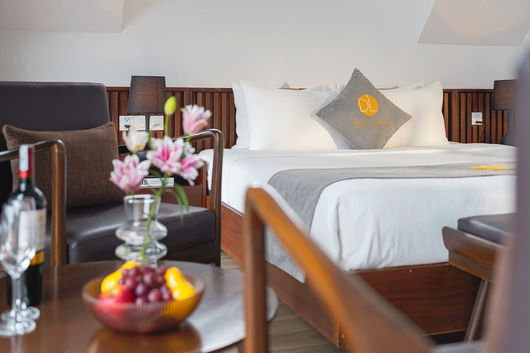 Quarto de hotel com cama, mesa de cabeceira com uma fruteira e uma garrafa, uma cadeira e um abajur.