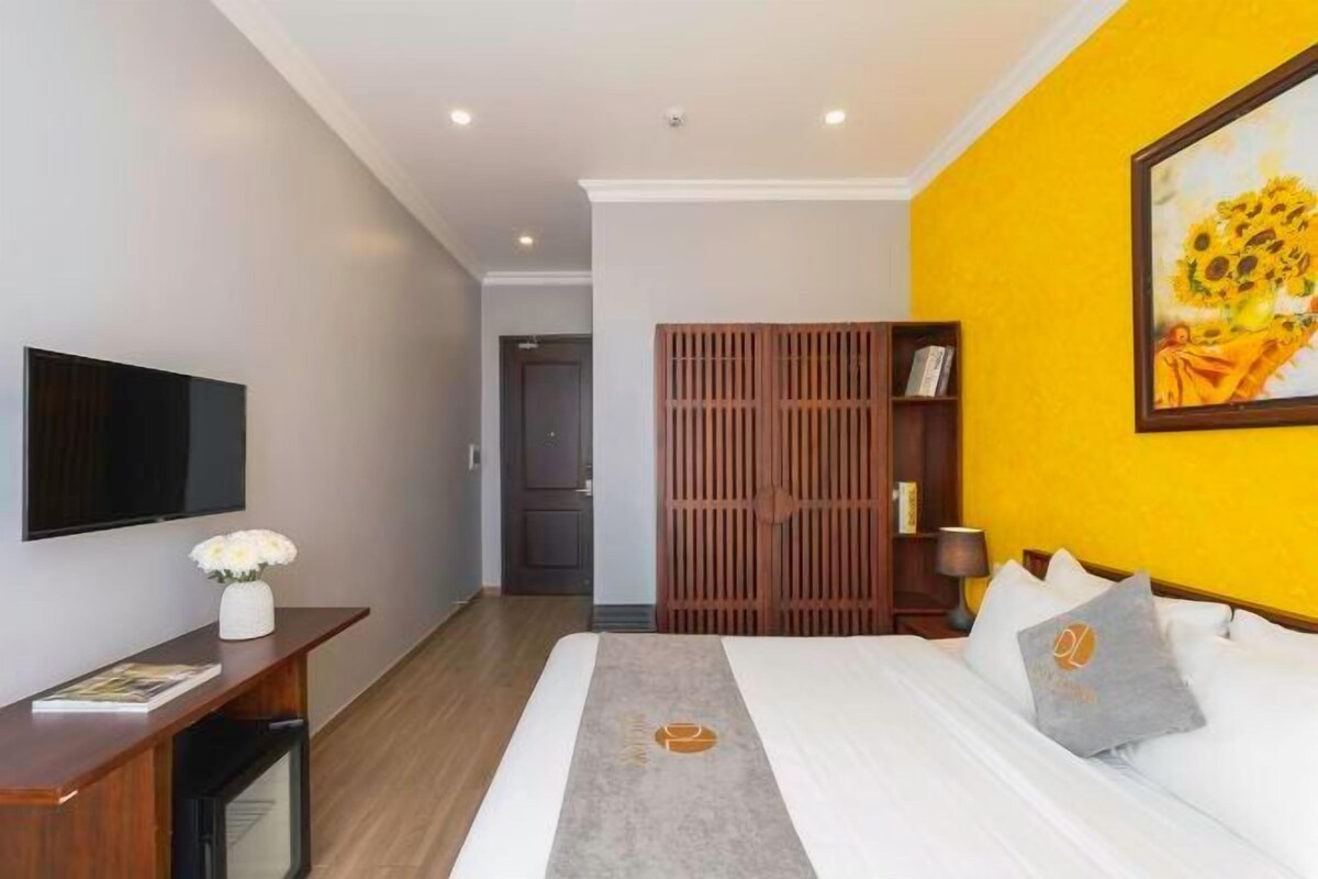 A hotel room with a bed, a flat-screen TV, a desk with a vase of flowers, a bookshelf, and a painting on the wall.
