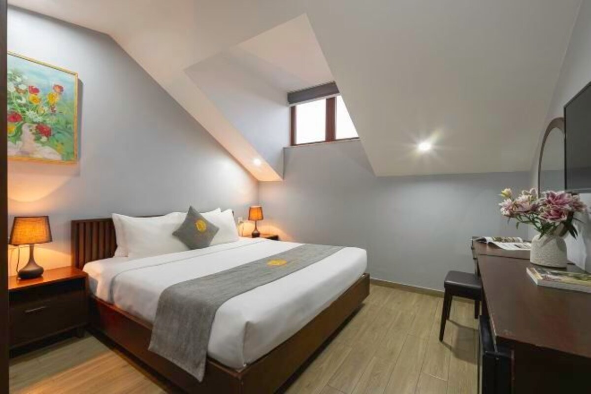 A modern bedroom with a skylight, a bed with a gray blanket, two bedside lamps, a desk with a vase of flowers, and a painting on the wall.