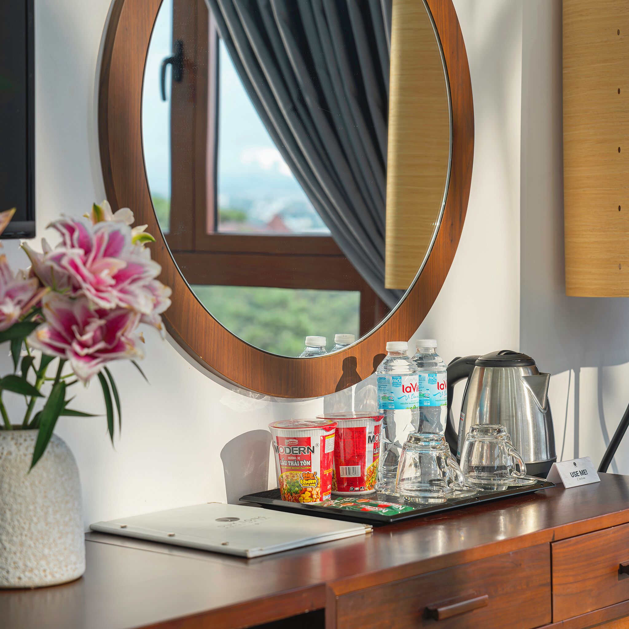 A hotel room with a wooden desk, a mirror, a vase of flowers, a laptop, a tray with a kettle, water bottles, and a coffee maker.