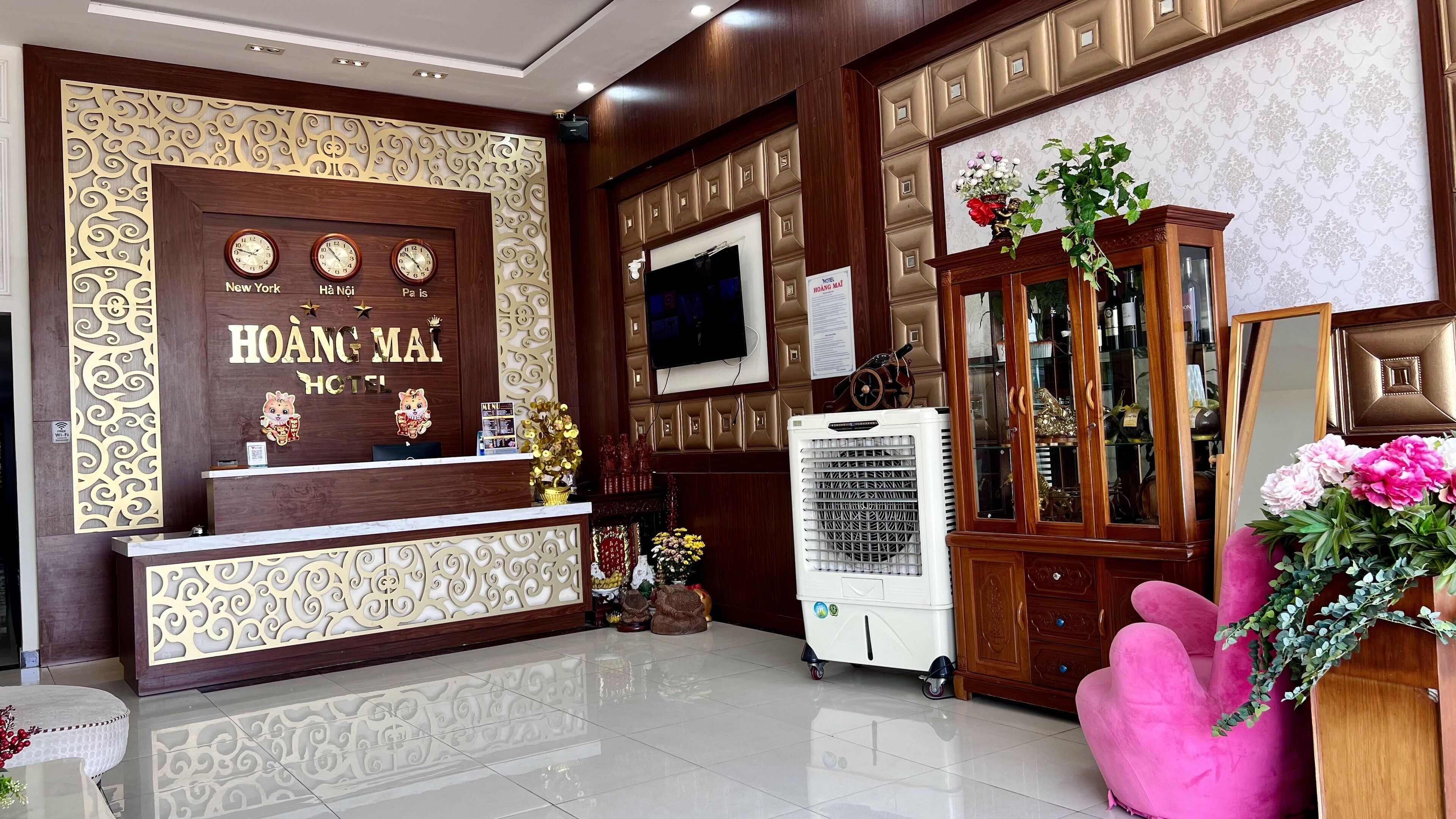 A hotel reception area with a wooden reception desk, a television, a cabinet with glass doors, and a pink chair.