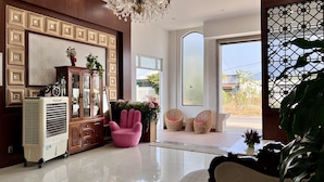 Una habitación amplia con un ventanal grande, un aparador de madera con puertas de cristal, un sillón rosa, sillas de mimbre y una planta en maceta.