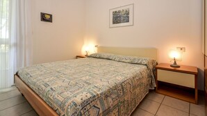 A neatly made bed with patterned bedding, two bedside tables with lamps, a framed picture on the wall, and a window with curtains. - Nice apartment for 4 guests with A/C, pool, TV and terrace (Bibione)