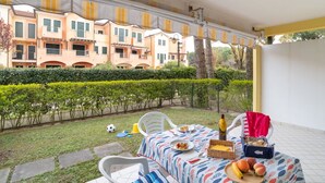 An outdoor patio with a table set for a meal, surrounded by a hedge and a row of apartment buildings in the background. - Nice apartment for 4 guests with A/C, pool, TV and terrace (Bibione)