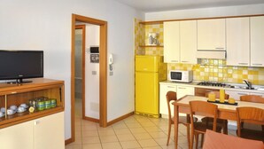 A compact kitchen with a microwave, stove, and refrigerator. There is a dining table with chairs and a television on a shelf. - Nice apartment for 4 guests with A/C, pool, TV and terrace (Bibione)