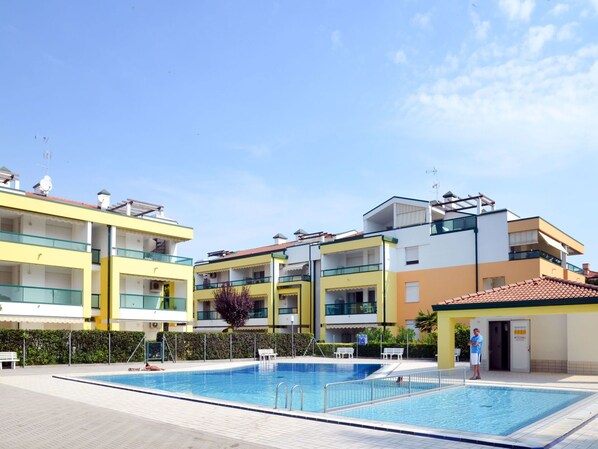 A modern hotel with a swimming pool, multiple balconies, and a clear blue sky.