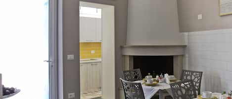 A dining area with a table set for four, chairs with intricate back designs, a white tiled wall, and a visible kitchen area in the background.