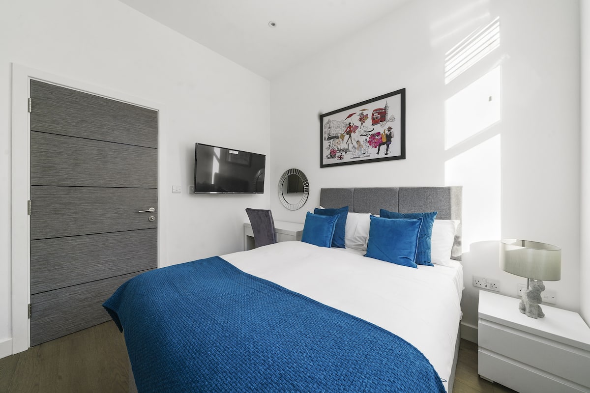 A modern bedroom with a large bed, a flat-screen TV, a framed artwork, and a bedside table with a lamp.