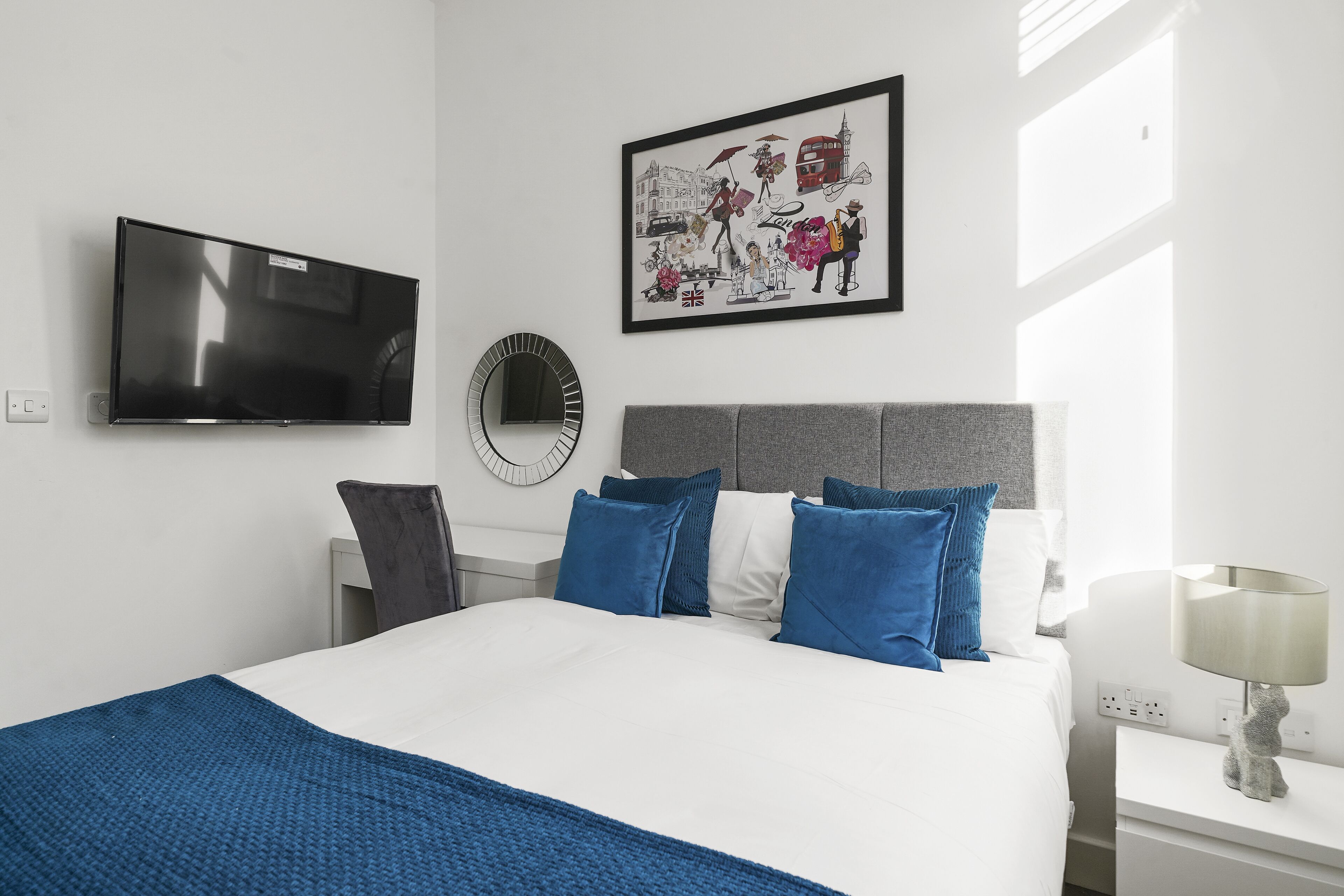 A modern hotel room with a large bed, a flat-screen TV, a framed cityscape artwork, and a bedside table with a lamp.