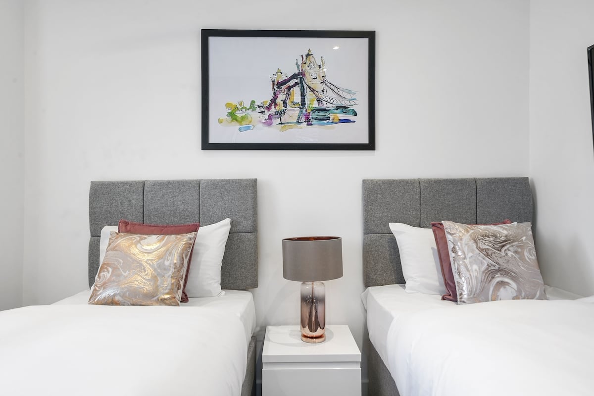 A hotel room with two beds, grey headboards, a white nightstand with a lamp, and a framed picture of a bridge on the wall.
