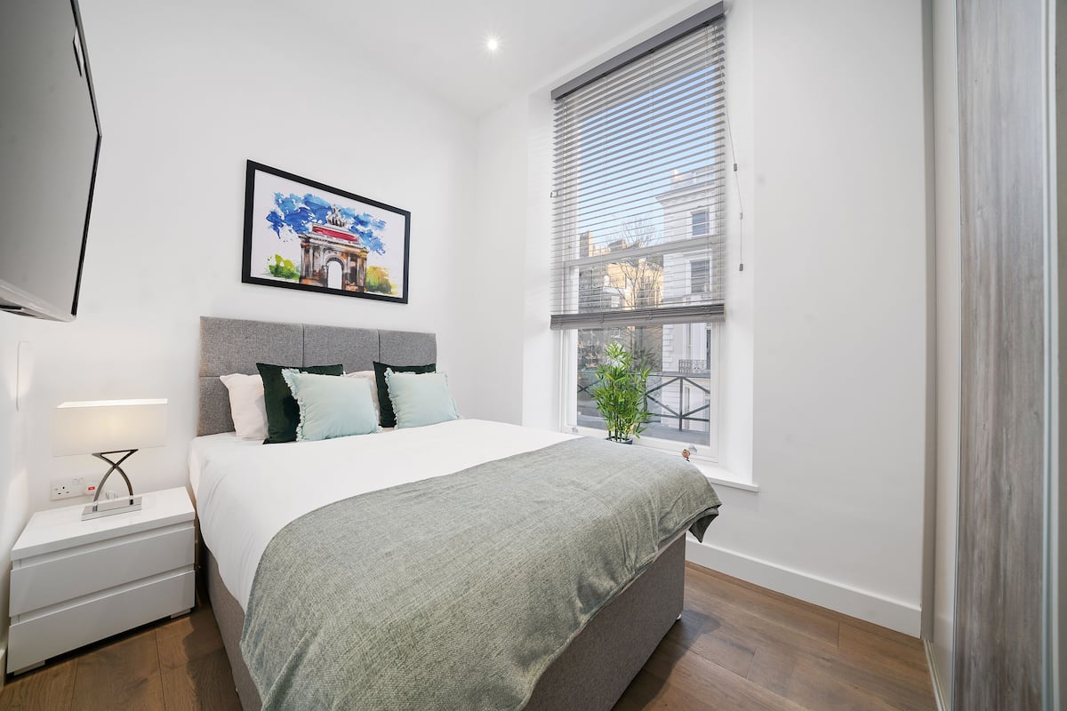 A modern bedroom with a bed, bedside table, and a window with blinds.