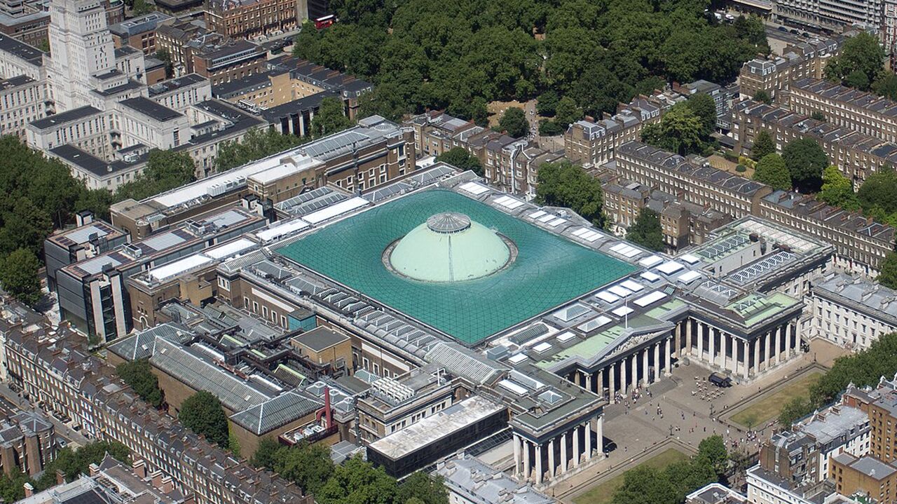 Vista aérea de una gran plaza con un área circular central, rodeada de edificios históricos y una estructura moderna con techo verde.
