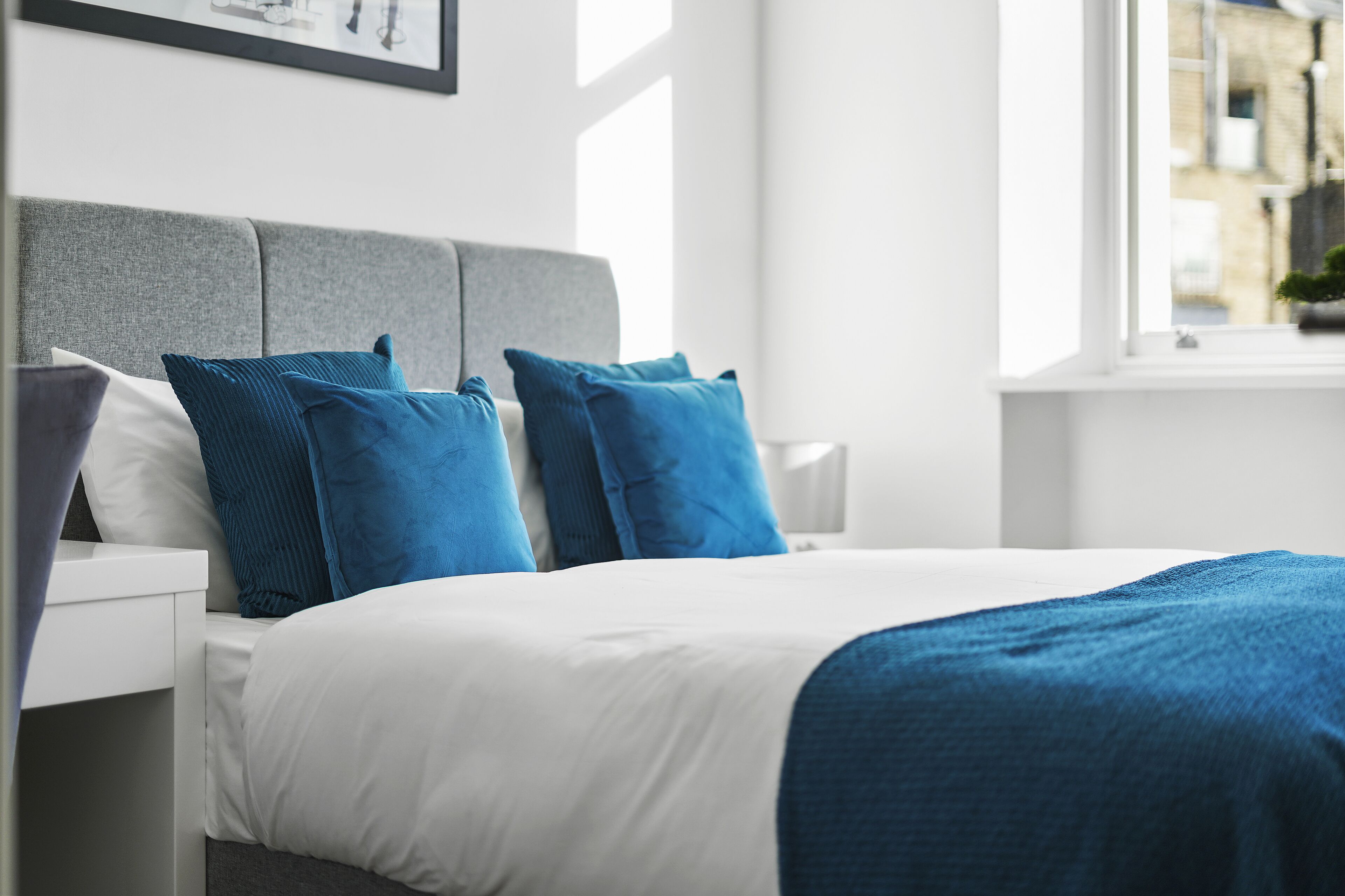 A neatly made bed with white and blue bedding, a grey headboard, and a window with a view of buildings.