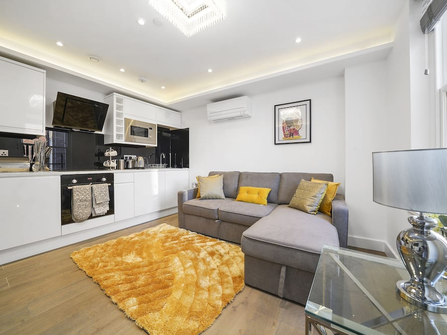 A modern kitchen with white cabinets, a flat-screen TV, and a grey sofa with yellow cushions.