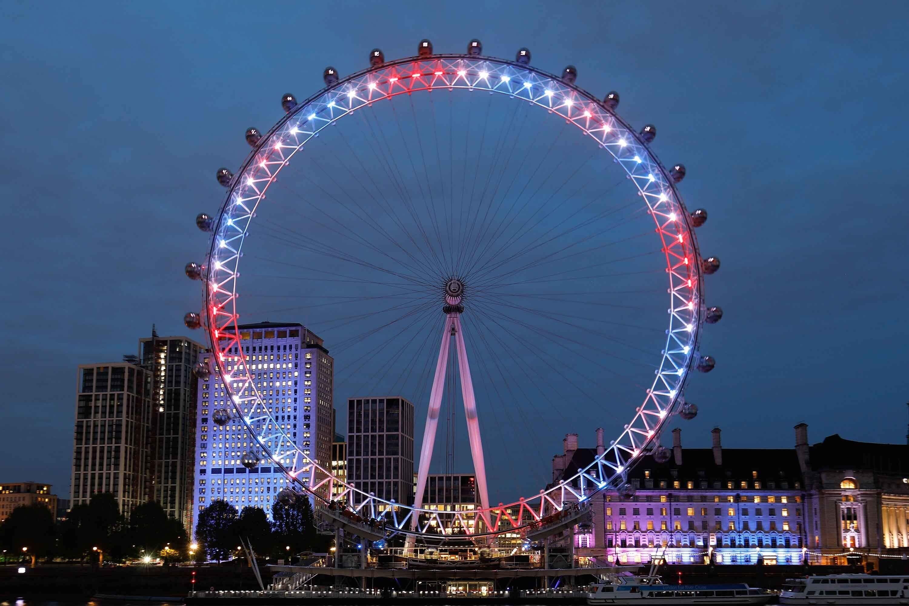 A large Ferris wheel with a red and white light pattern, surrounded by buildings and a river.