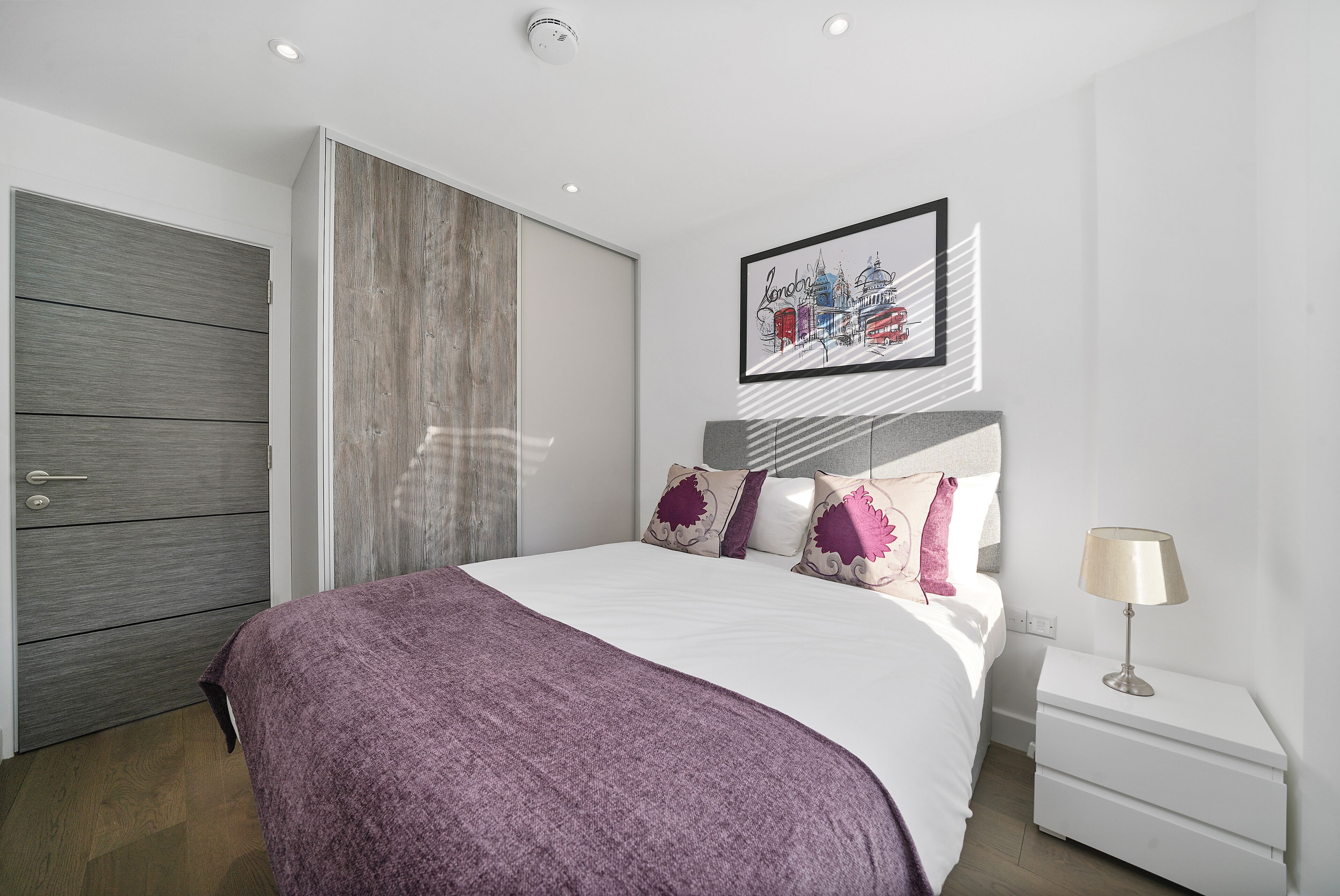 A modern bedroom with a large bed, a grey wardrobe, a nightstand with a lamp, and a framed cityscape artwork on the wall.