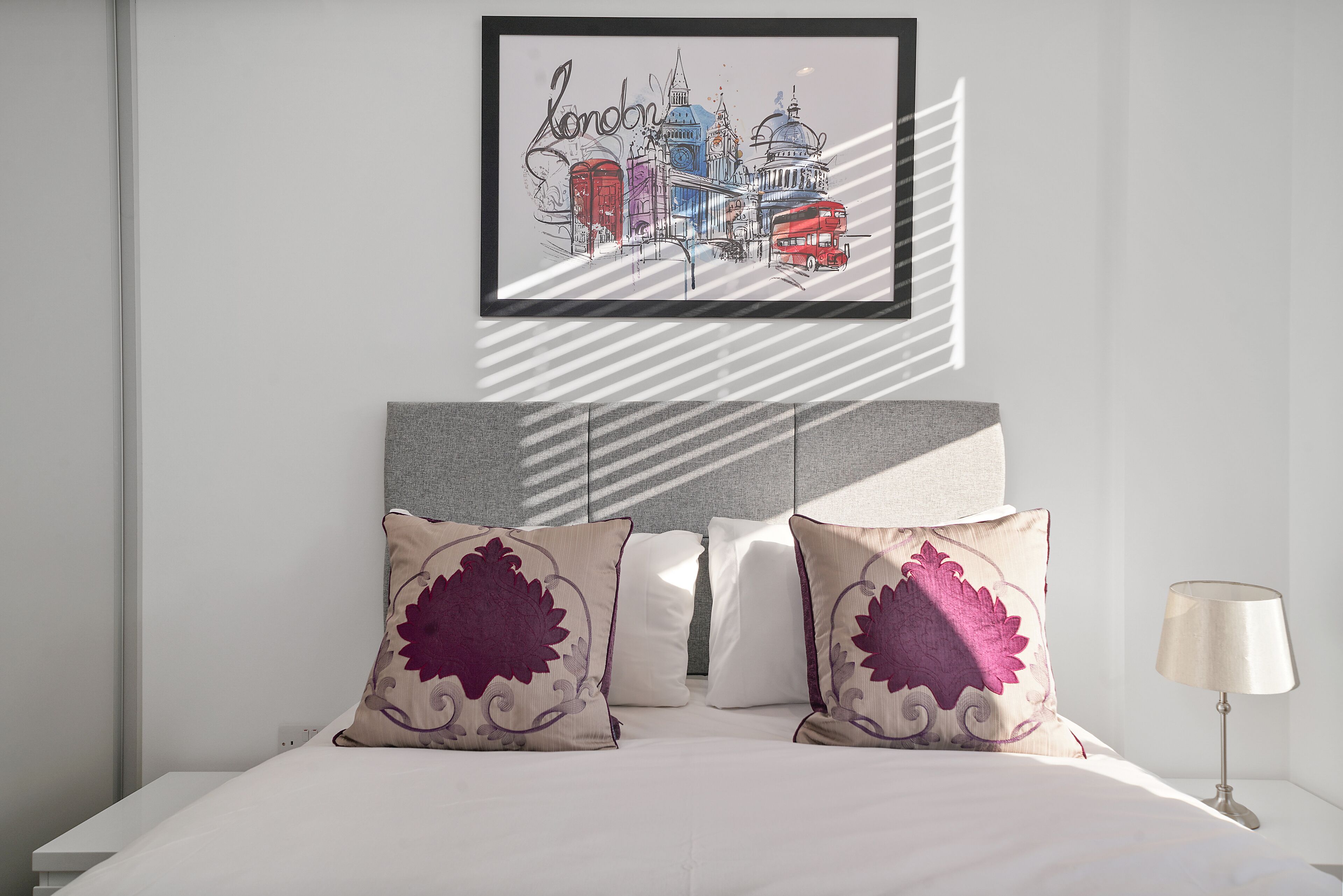 A neatly made bed with two decorative pillows, a framed artwork of London landmarks above it, and a bedside lamp.