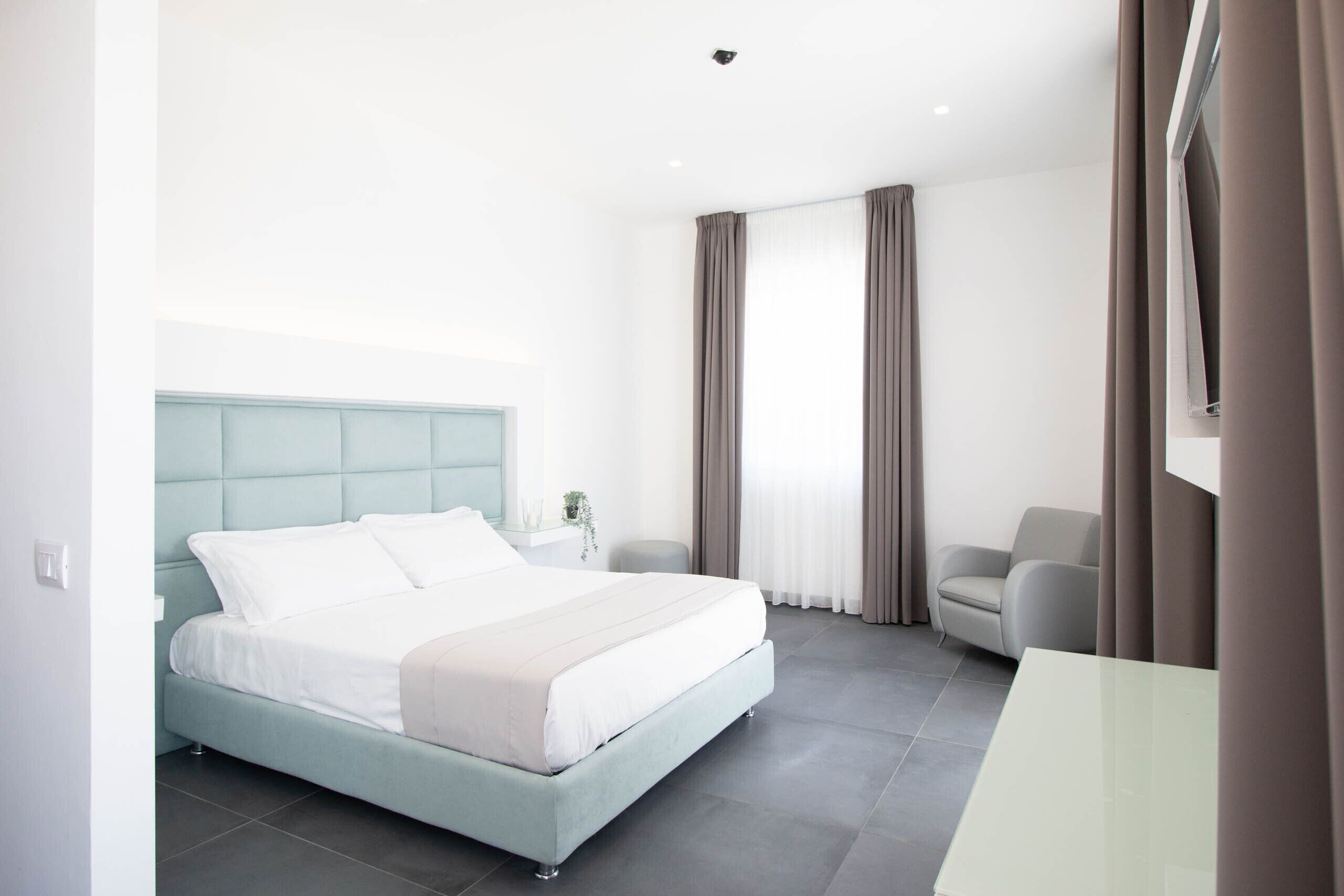 Una habitación de hotel moderna con una cama grande, una mesita de noche pequeña, una silla y un ventanal con cortinas.