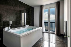 A modern bathroom with a large bathtub, dark marble walls, and a view of the sea through large windows. - Grand Hotel Serapide (Pozzuoli)