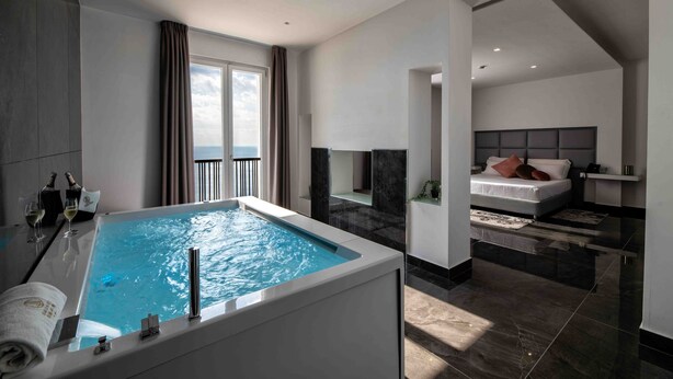 A modern hotel room with a large bathtub, a balcony with a view, and a separate bedroom with a bed and nightstand. - Grand Hotel Serapide (Pozzuoli)