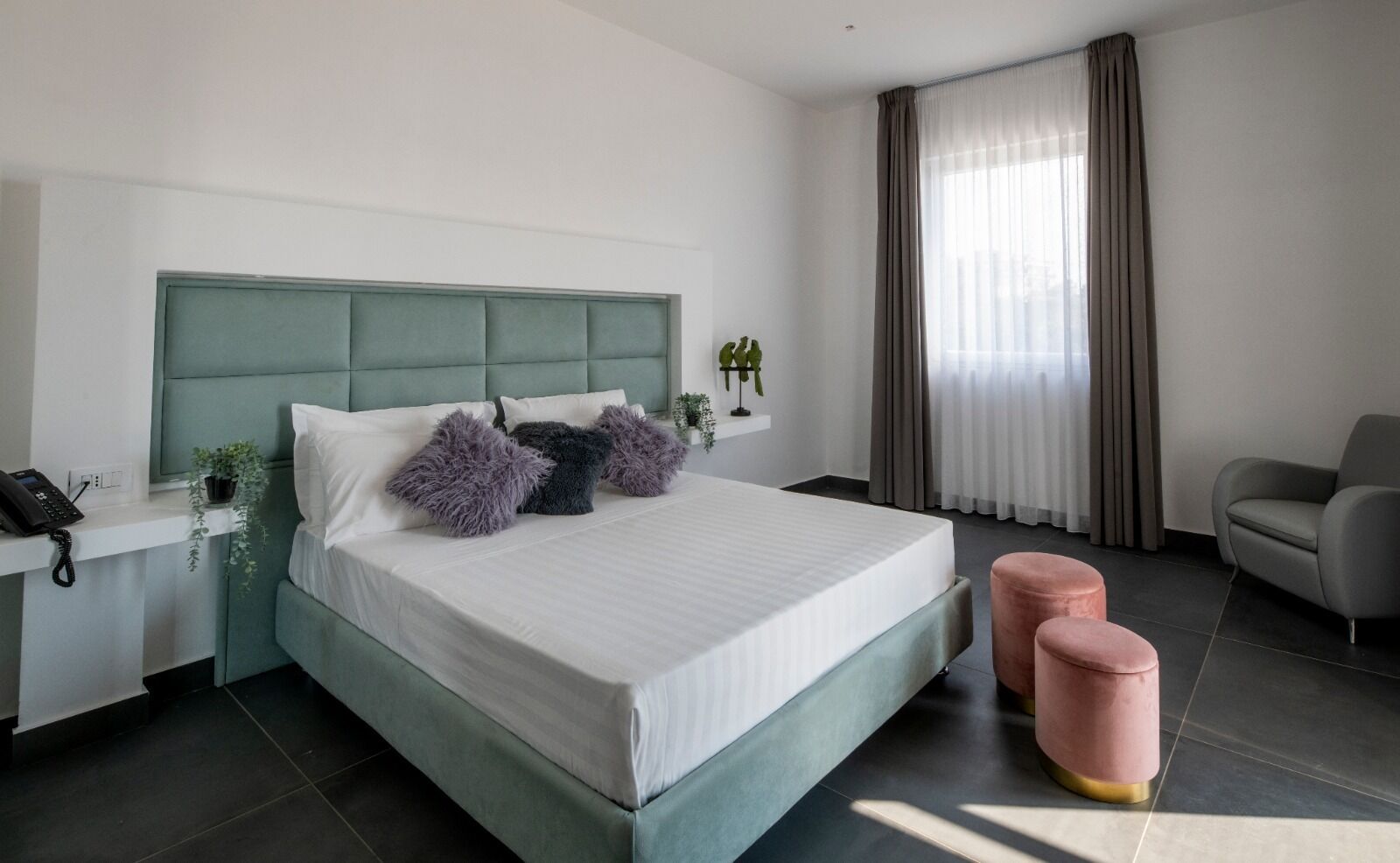 Ein modernes Hotelzimmer mit einem großen Bett, einem Nachttisch mit Telefon, einem Sessel und einem Schreibtisch mit Computer.