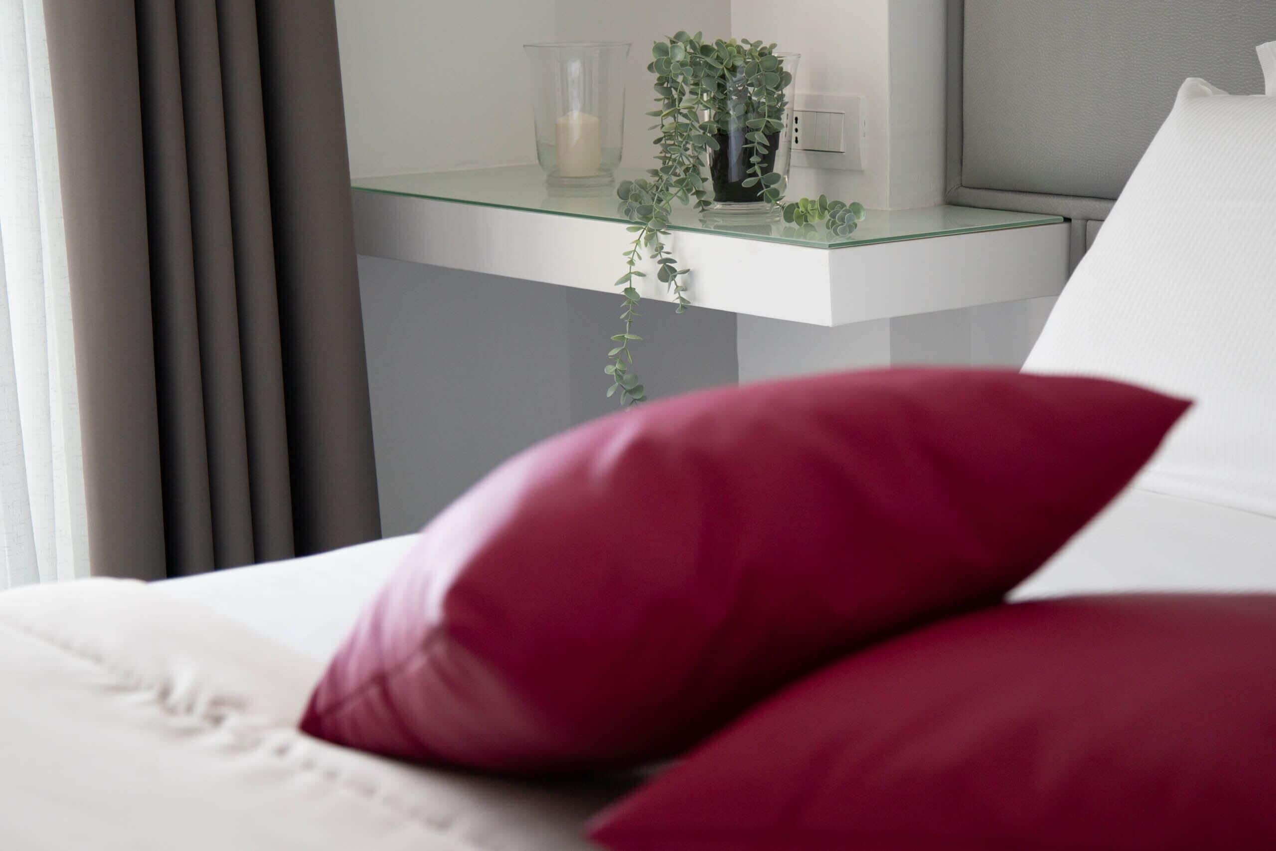 Een bed met rode kussens, een glazen bijzettafeltje met een kaars en een plant, en een wit hoofdeinde.