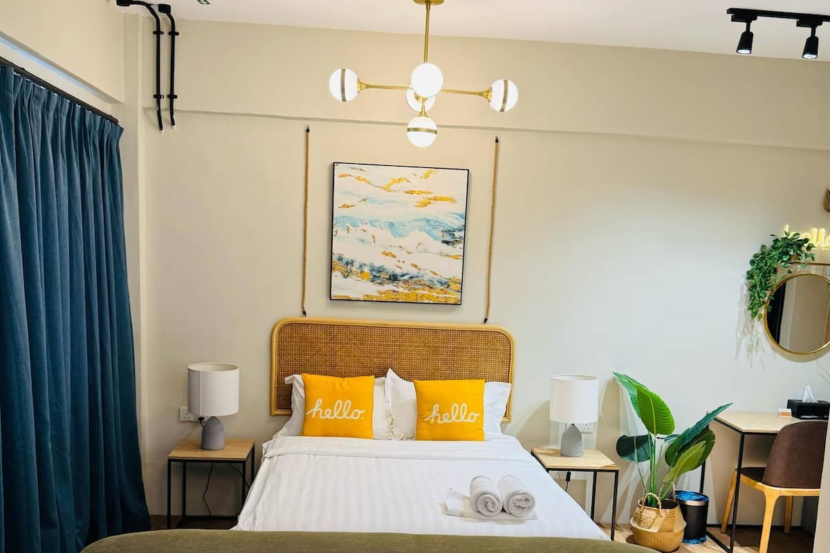 A hotel room with a large bed, two bedside tables, a desk, a chair, a sofa, a ceiling light fixture, a wall-mounted plant, and a painting.