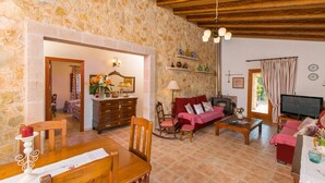 A cozy living room with a stone wall, wooden ceiling, a fireplace, a television, and a dining area with a table and chairs. - SA ROTA Finca for 6 people in Ariany with pool (Ariany)