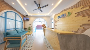 A reception area with a wooden reception desk, a blue sofa, a small table with a plant, and a TV mounted on the wall. - TURI STAYpoint (Nghia Lo)