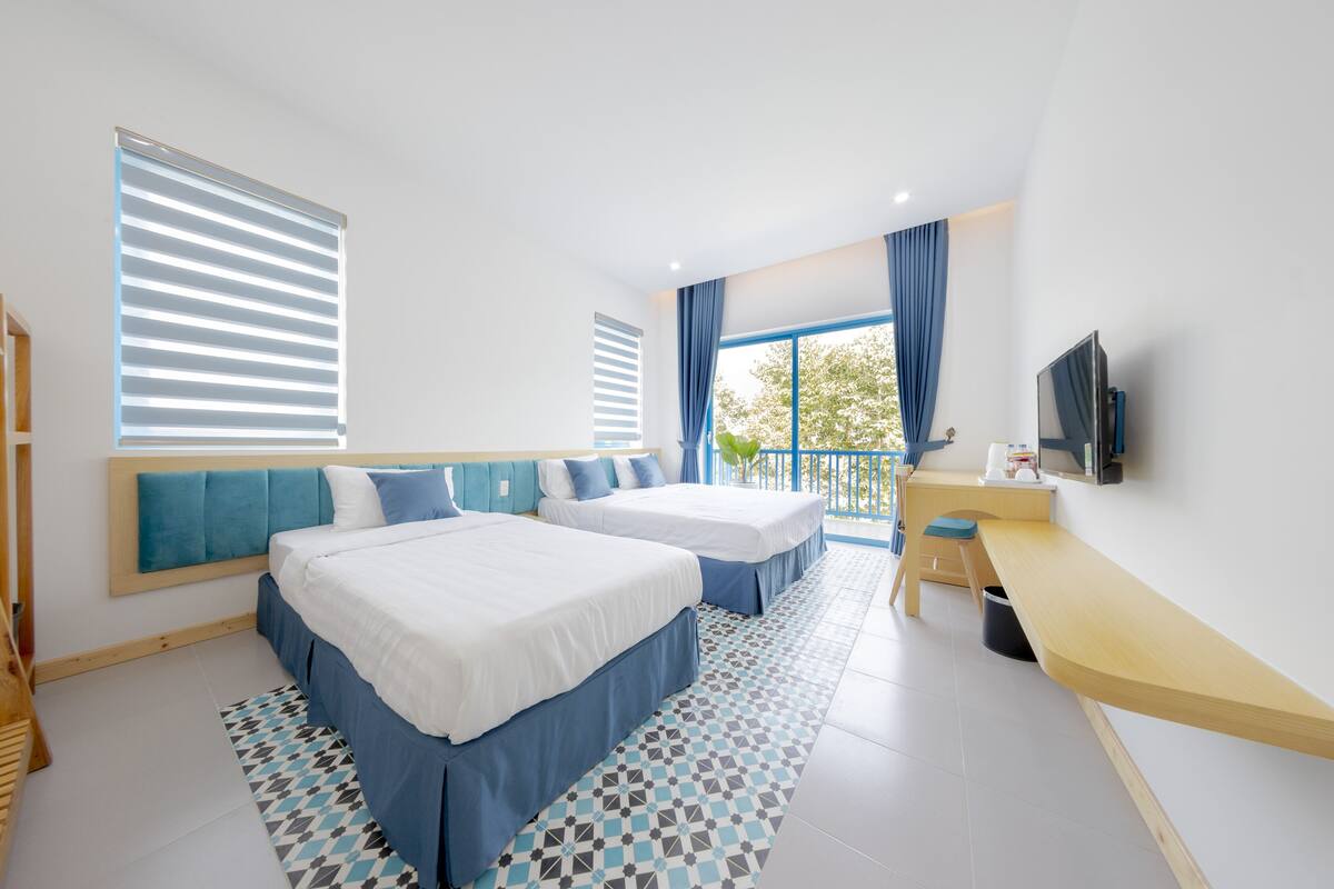 A modern hotel room with two beds, a desk, a TV, and a balcony with blue curtains.