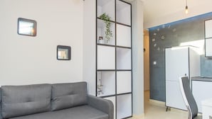 En moderne stue med en grå sofa, et hvidt spisebord og et køkkenområde med et køleskab og skabe.