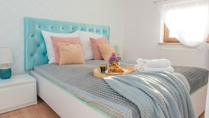 En pænt redt seng med et blåt sengegærde, et natbord med en lampe, og et bakke med croissanter og drikke.