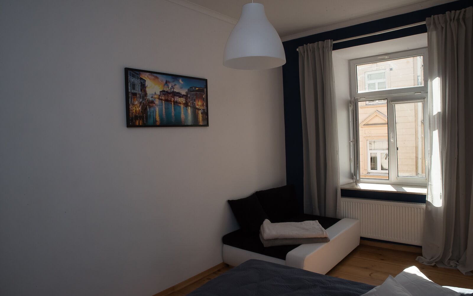 Una stanza con un letto, una finestra con tende, un termosifone e un quadro che raffigura un panorama urbano.