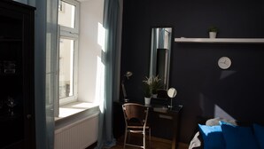Una habitació amb una finestra gran, un escriptori amb una cadira, una planta petita, un rellotge i un sofà blau.