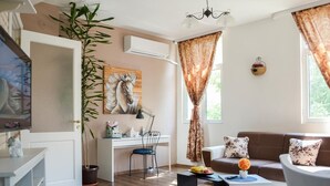 Un salon comprenant un canapé, une table basse et un bureau avec une chaise. On y trouve également une grande plante, un tableau et une fenêtre avec des rideaux.