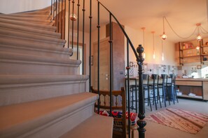 Una escalera con barandilla de madera y balaustres negros, que conduce a un comedor con taburetes altos y una cocina al fondo.