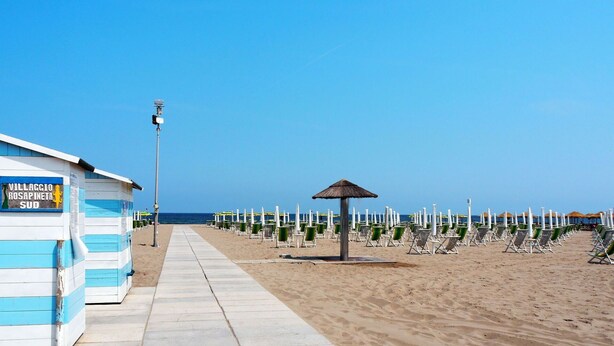 Una platja amb una filera de cabanes de bany i sombrerets, cel serè i un camí de sorra que condueix al mar.