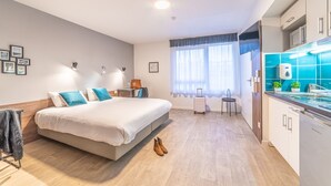 A modern hotel room with a large bed, a small desk, a window with curtains, and a kitchen area with blue tiles. - Break & Home Paris Italie Porte De Choisy (Ivry-sur-Seine)