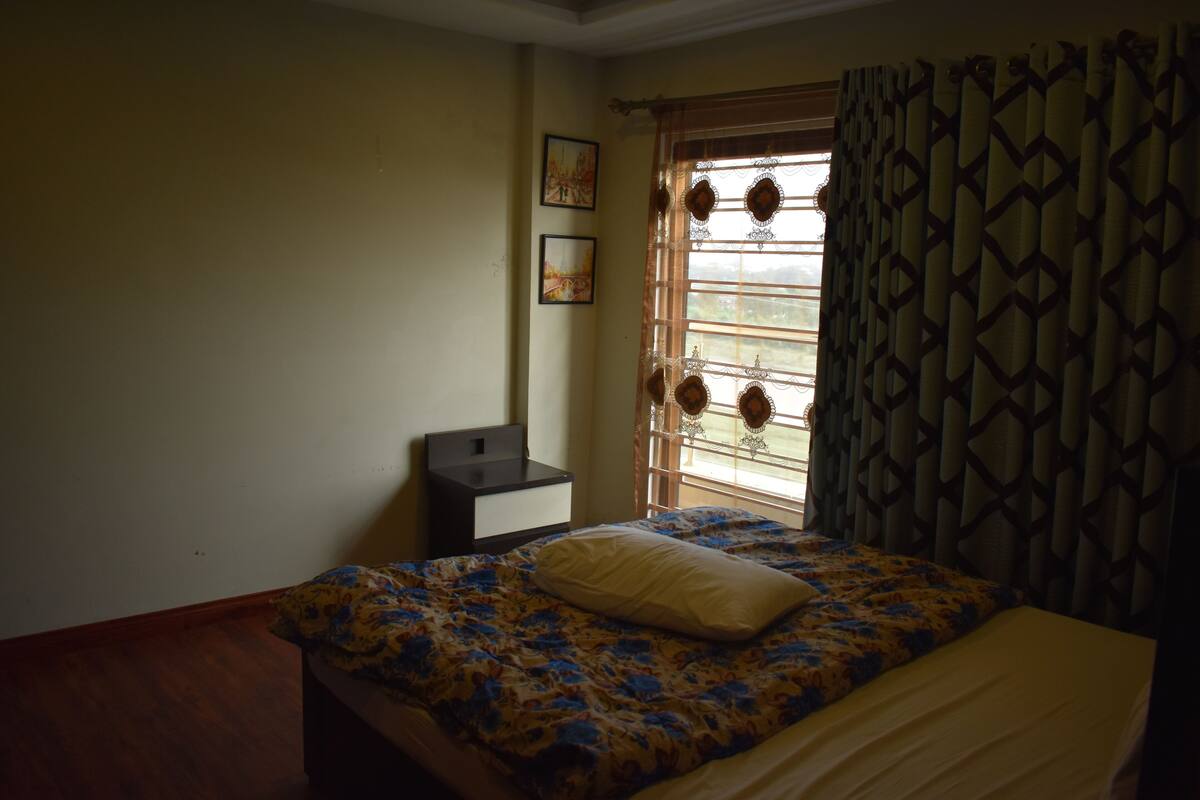 Un dormitorio con cama, mesita de noche, ventana con cortinas y un cuadro con dos marcos en la pared.