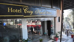L'ingresso di un hotel con un'ampia vetrata che riflette l'interno e un cartello con la scritta "Hotel Cozy Knock Coffee".