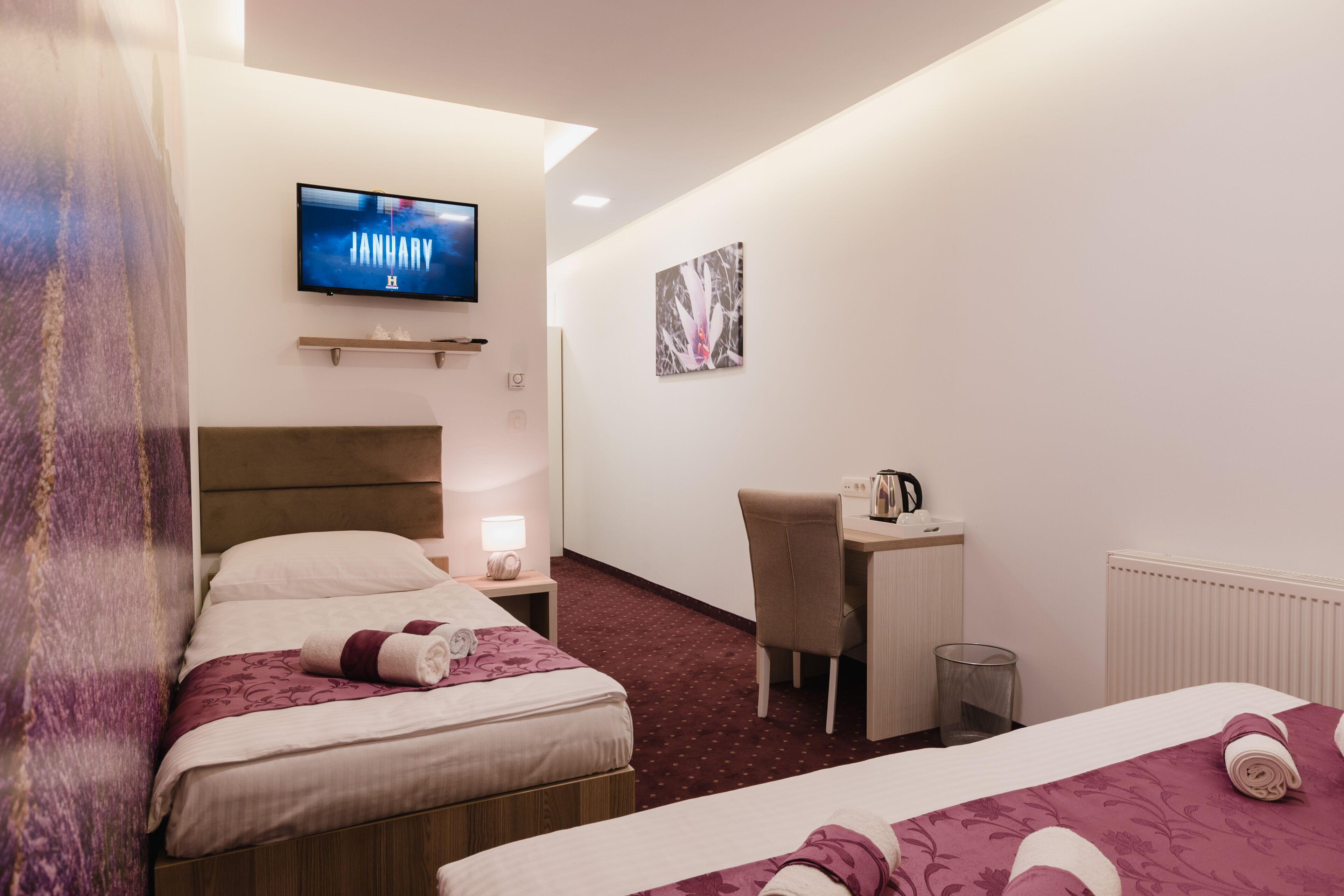 A hotel room with two beds, a desk, a chair, a television, and a radiator.