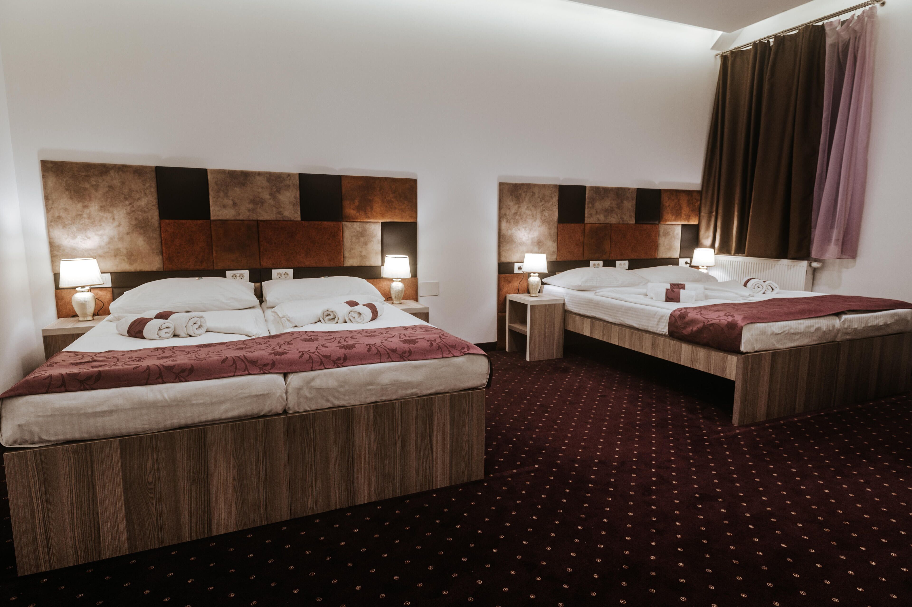 A hotel room with two beds, each with a wooden headboard, white linens, and a patterned throw. There are bedside tables with lamps and a large window with curtains.