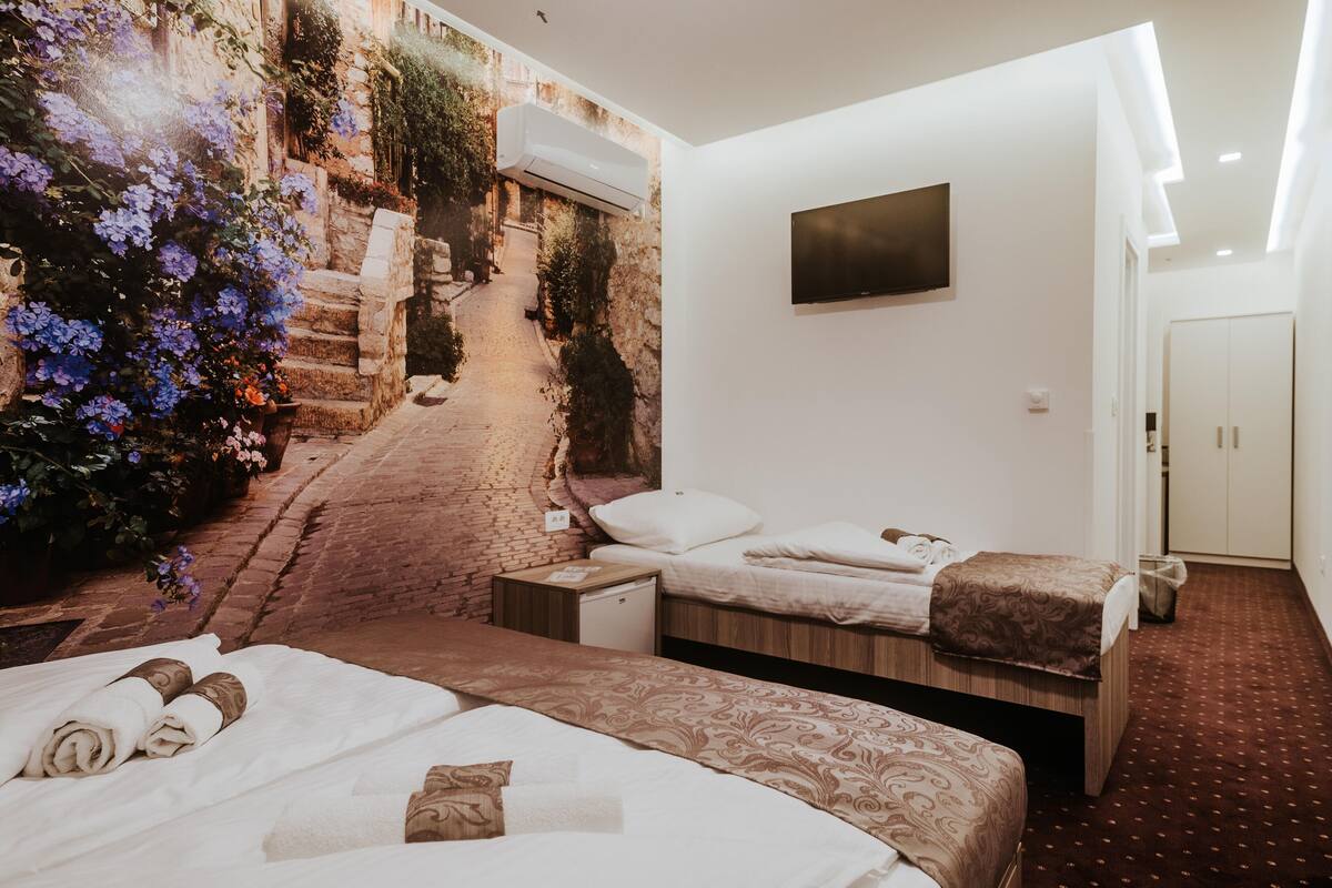 A hotel room with two beds, a flat-screen TV, a mural of a cobblestone street, and a small desk with a lamp.