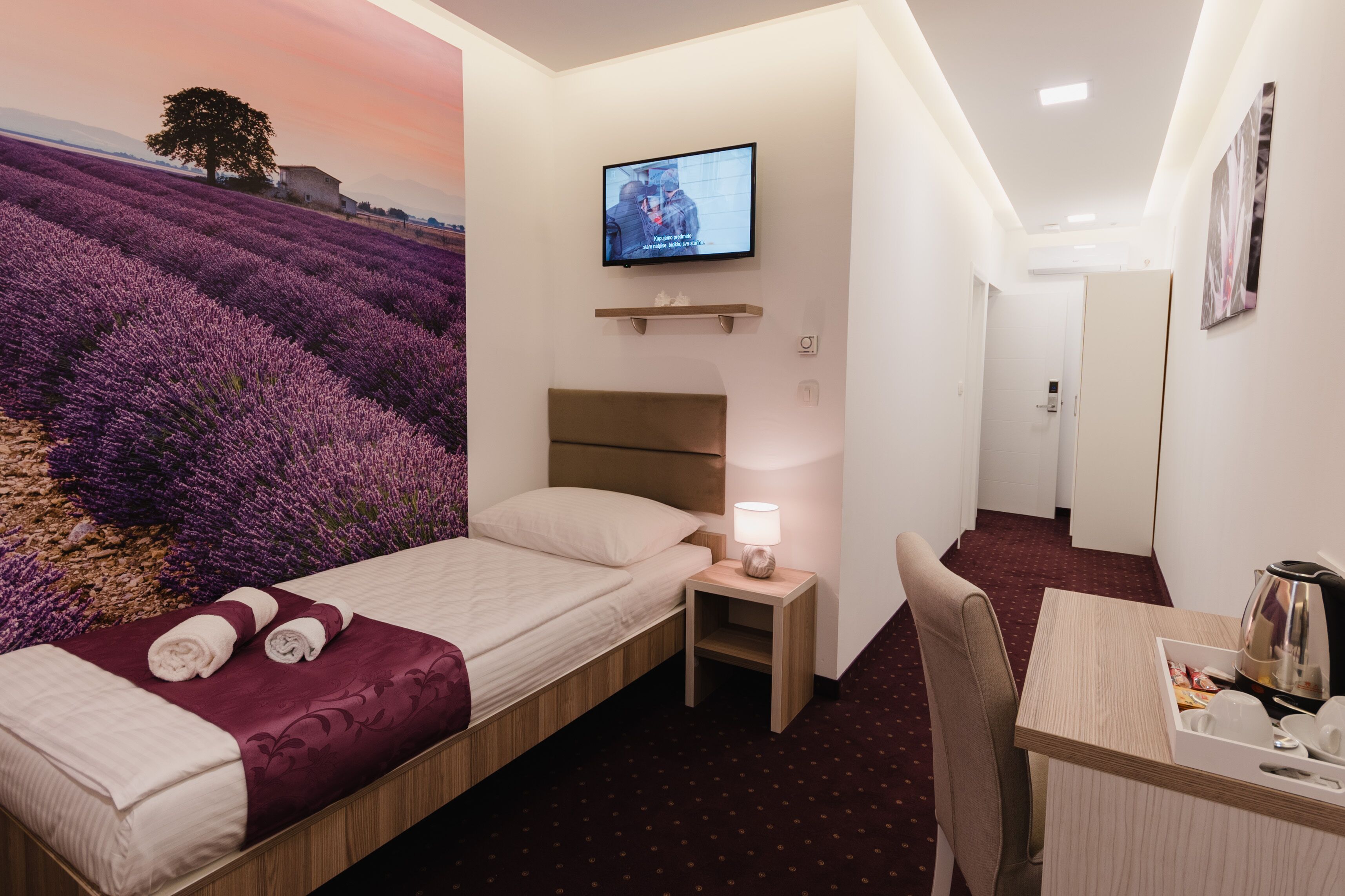 A hotel room with a bed, a desk, a TV, and a view of lavender fields.