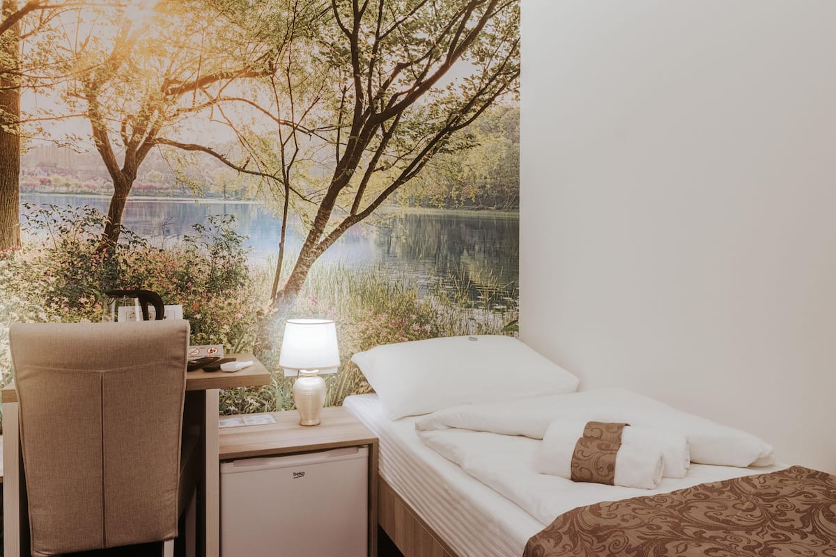 A hotel room with a bed, a desk with a lamp, a small refrigerator, and a chair.