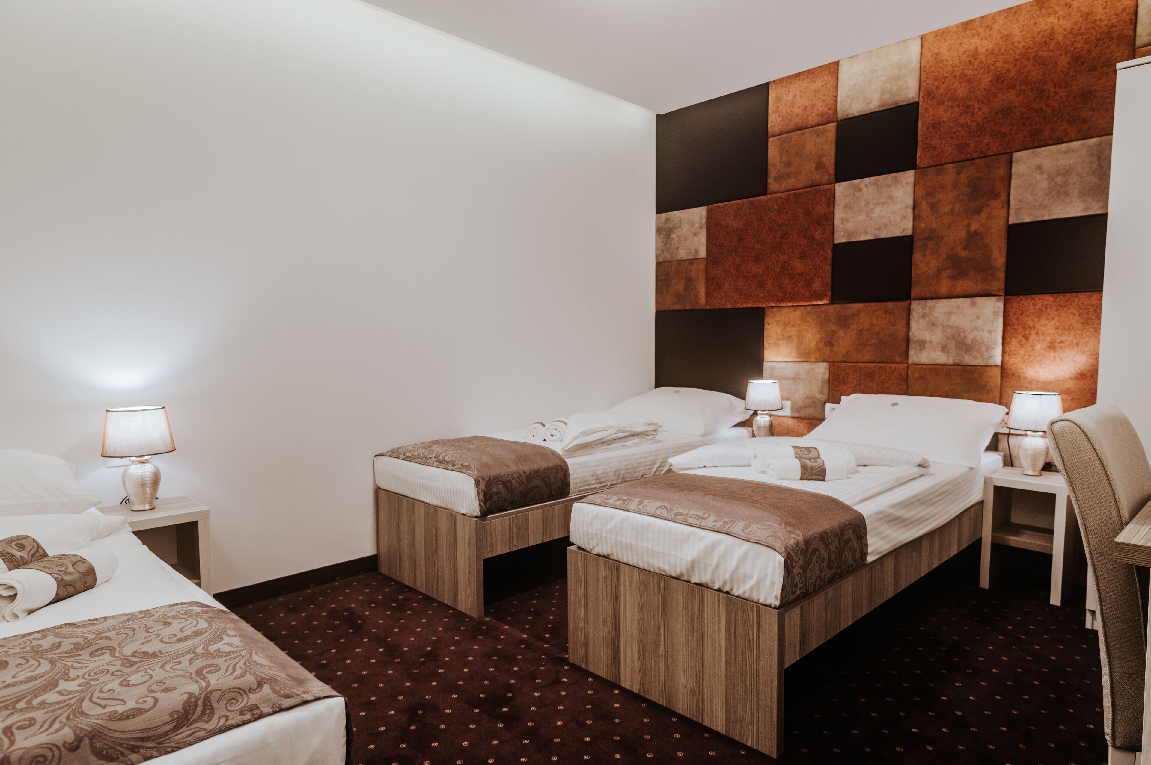 A hotel room with two single beds, a wooden headboard, a small table with a lamp, and a patterned carpet.