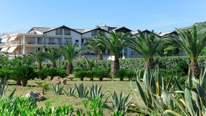 Un hotel de varios pisos con balcones, rodeado de un jardín ajardinado con diversas plantas y palmeras.
