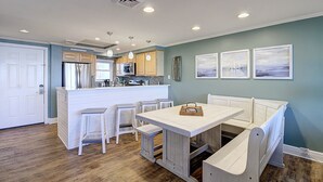 A kitchen with a central island, white bar stools, a dining table with benches, and a view of the ocean through the windows. - Ocean View Delight, Resort Pools, Private Boardwalk | MP301 (Port Aransas)