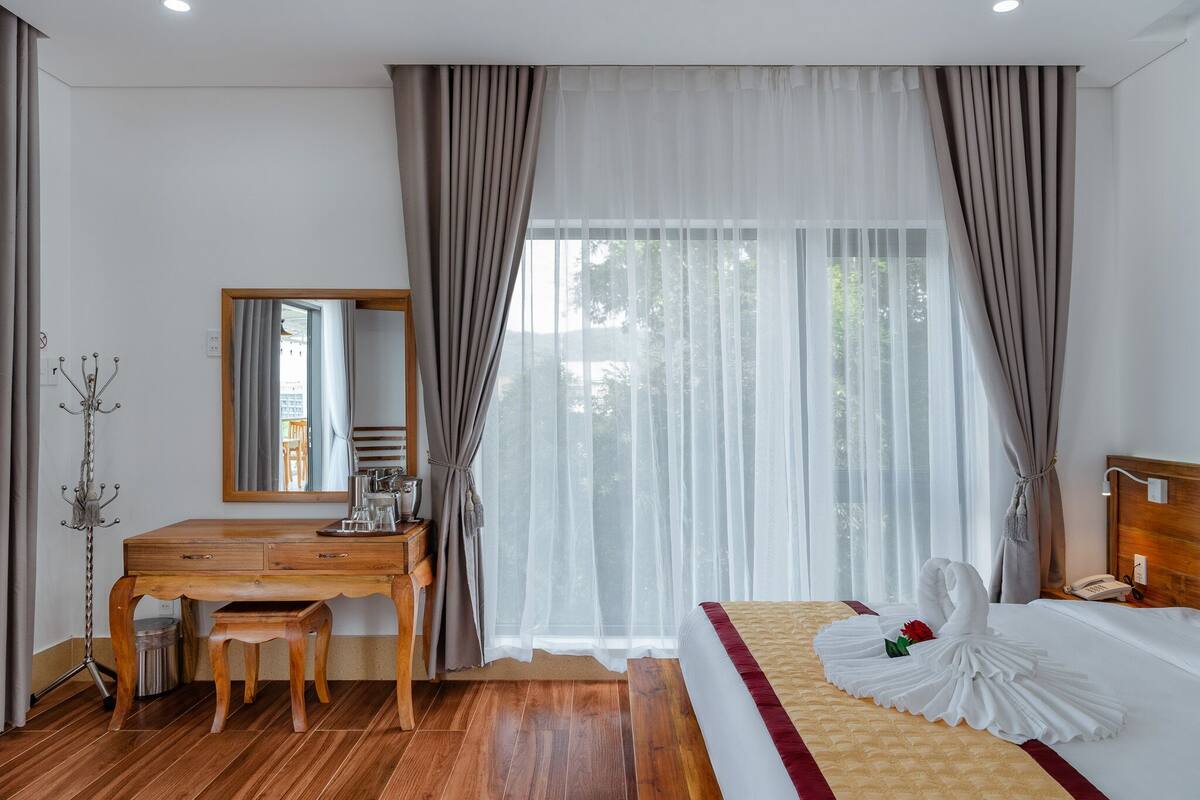 Ein Hotelzimmer mit einem Holztisch, einem Bett mit weißer Daunendecke und einer dekorativen Schwanenfigur, sowie einem großen Fenster mit transparenten Vorhängen.